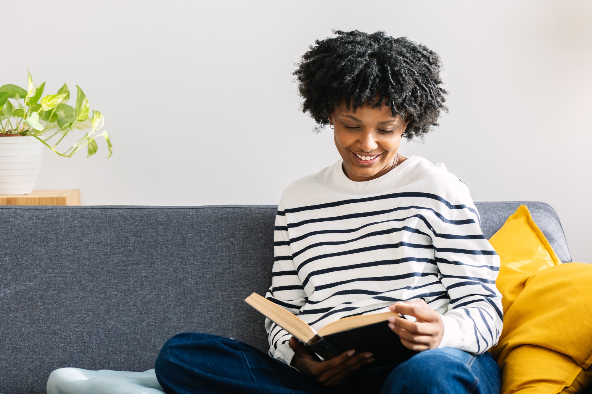 Happy beautiful African American woman reading a book sitting on sofa at home